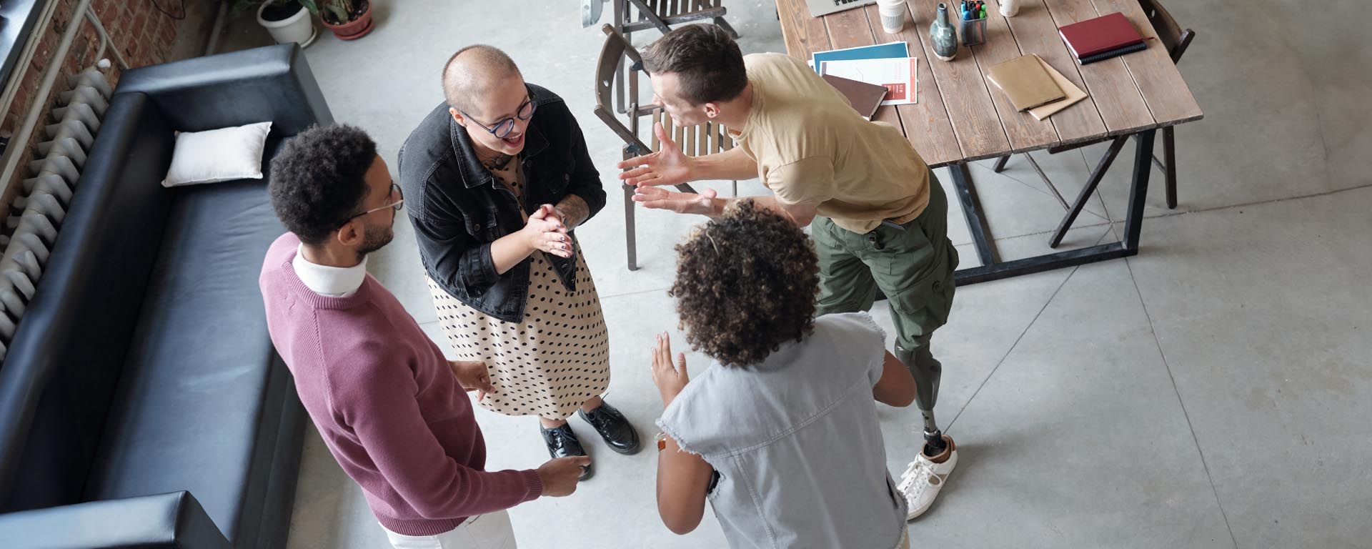 4 employees are facing each other in a team-building environment. One male employee has a prosthetic leg. One female has a shaved head implying recent chemotherapy treatment.