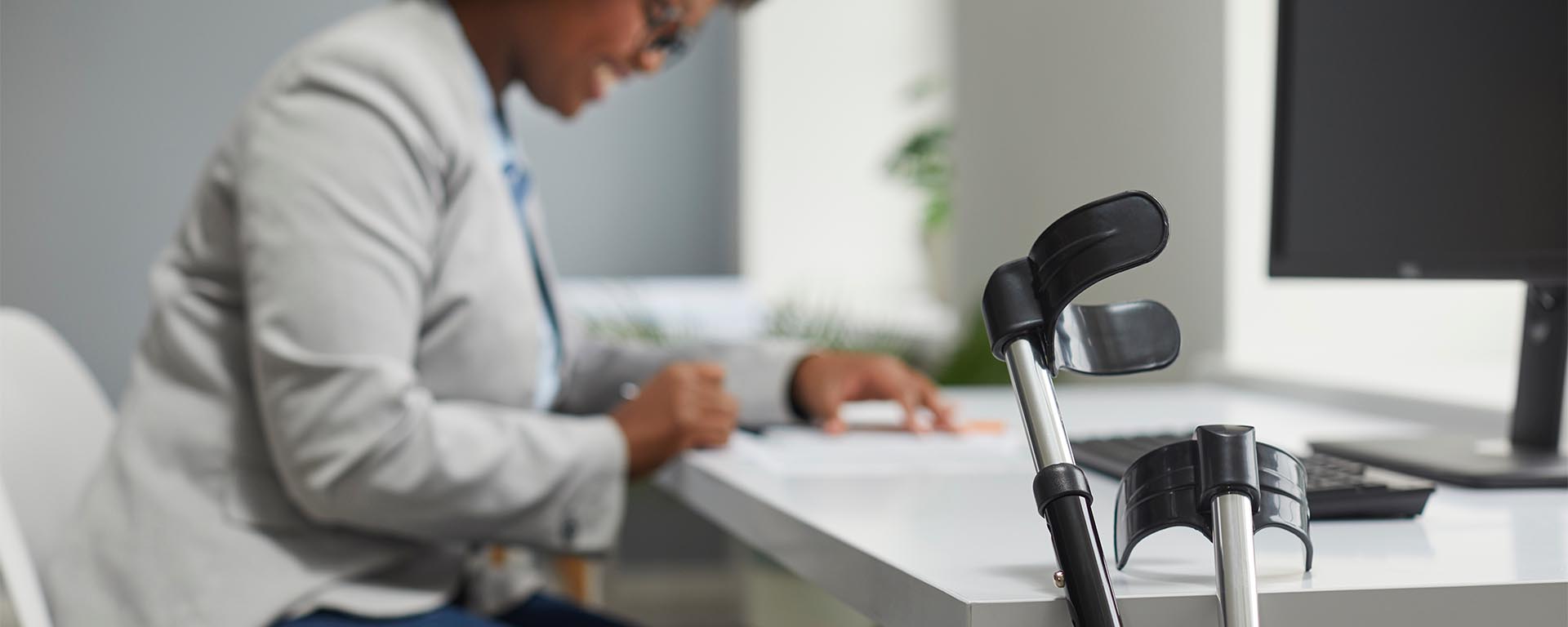 Focus on forearm crutches leaning against a work desk, with a blurred vision of a female employee in background.