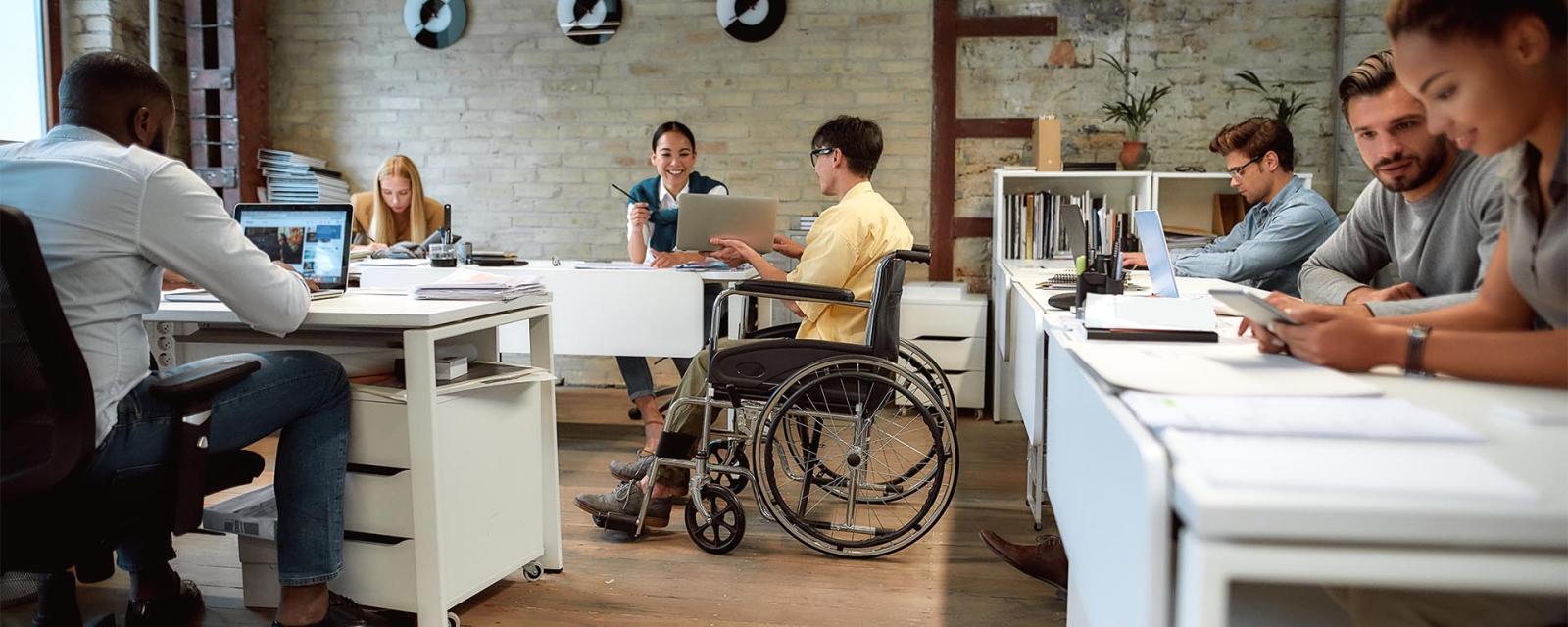 Office setting with disabled male employee in wheelchair presenting to female co-worker.