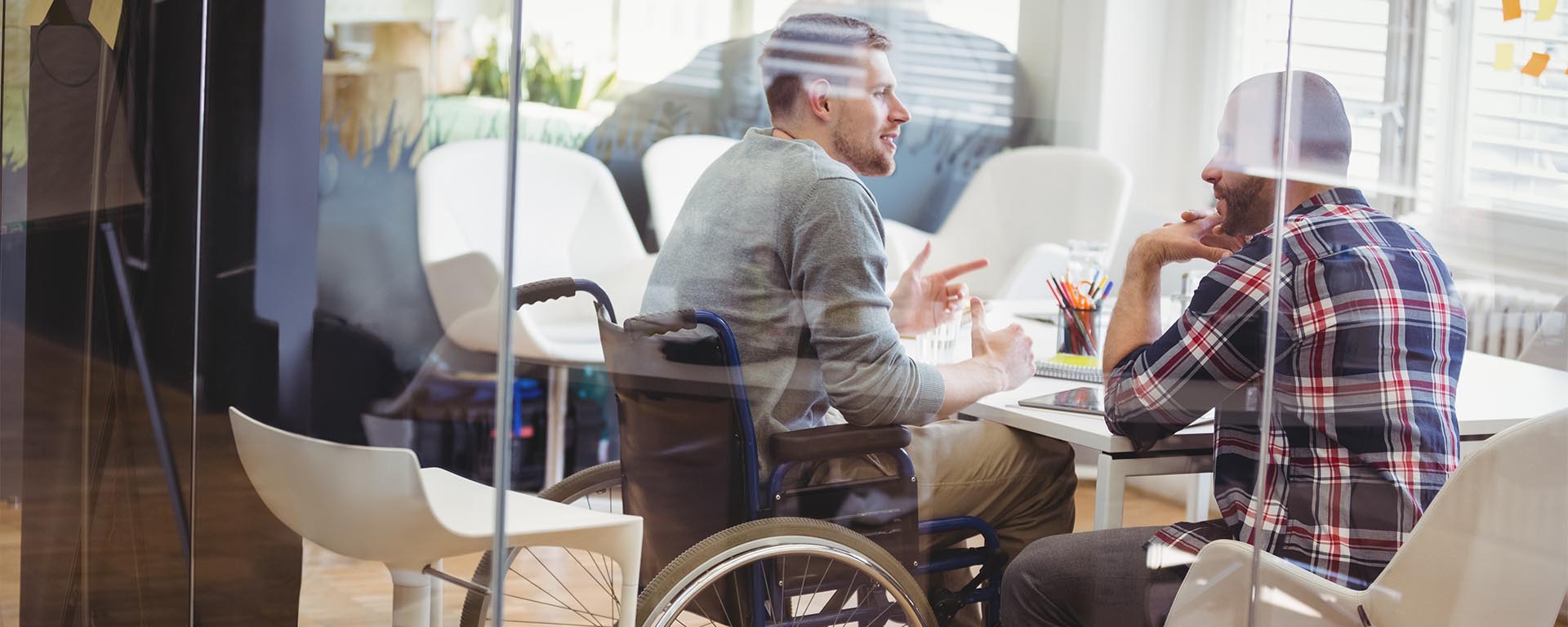 Two male co-workers are collaborating in a conference room. One disabled male is in a wheelchair.