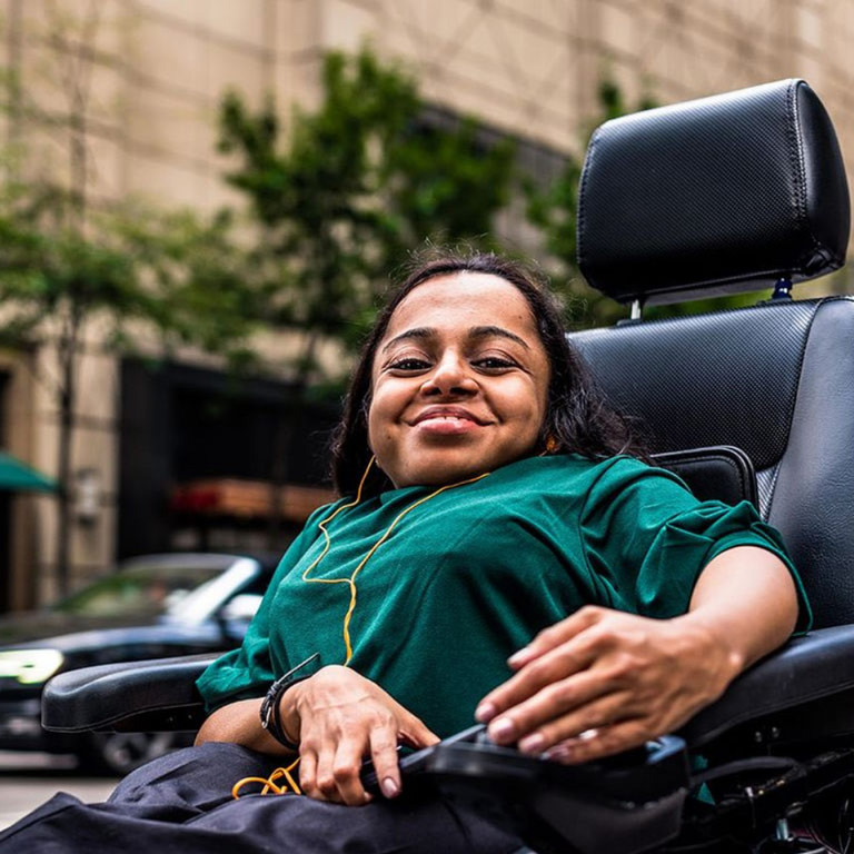 Disabled woman in motorized wheelchair on sidewalk smiling.