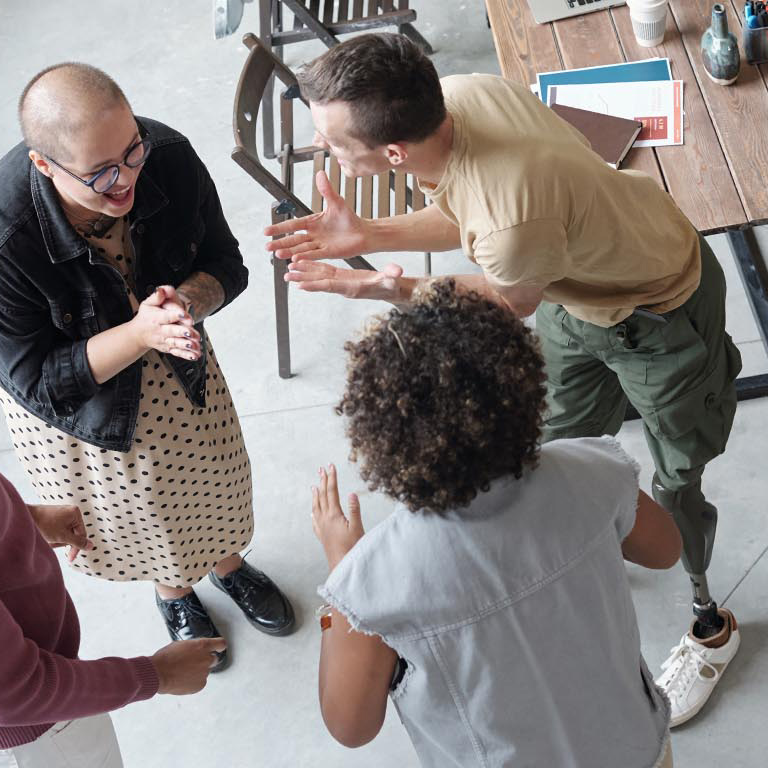 4 employees are facing each other in a team-building environment. One male employee has a prosthetic leg. One female has a shaved head implying recent chemotherapy treatment.