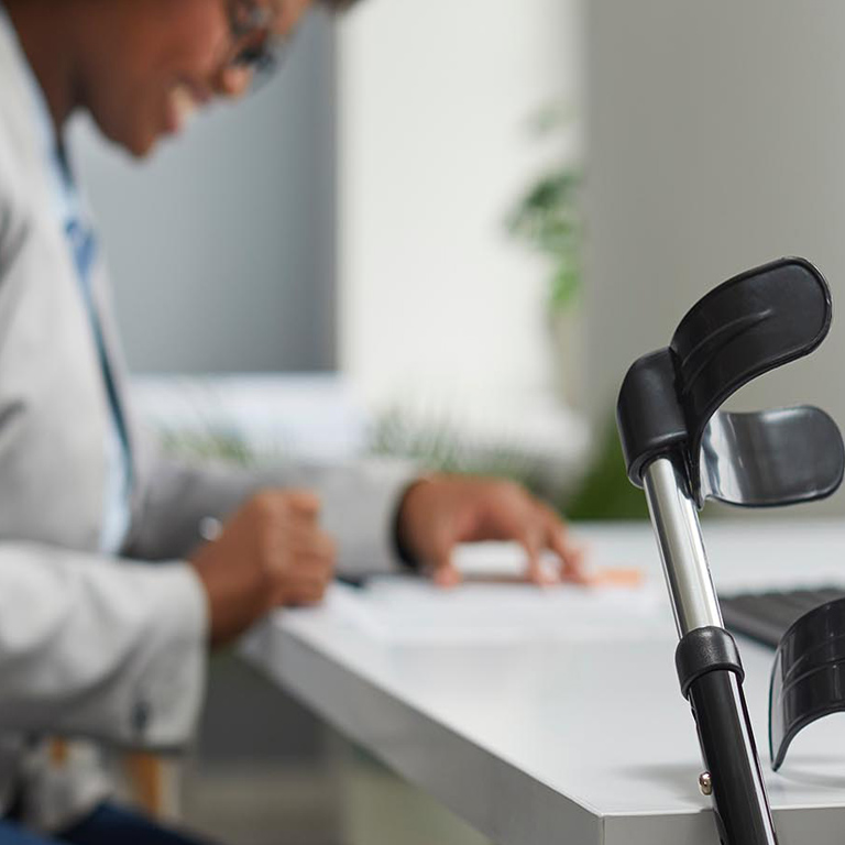 Focus on forearm crutches leaning against a work desk, with a blurred vision of a female employee in background.