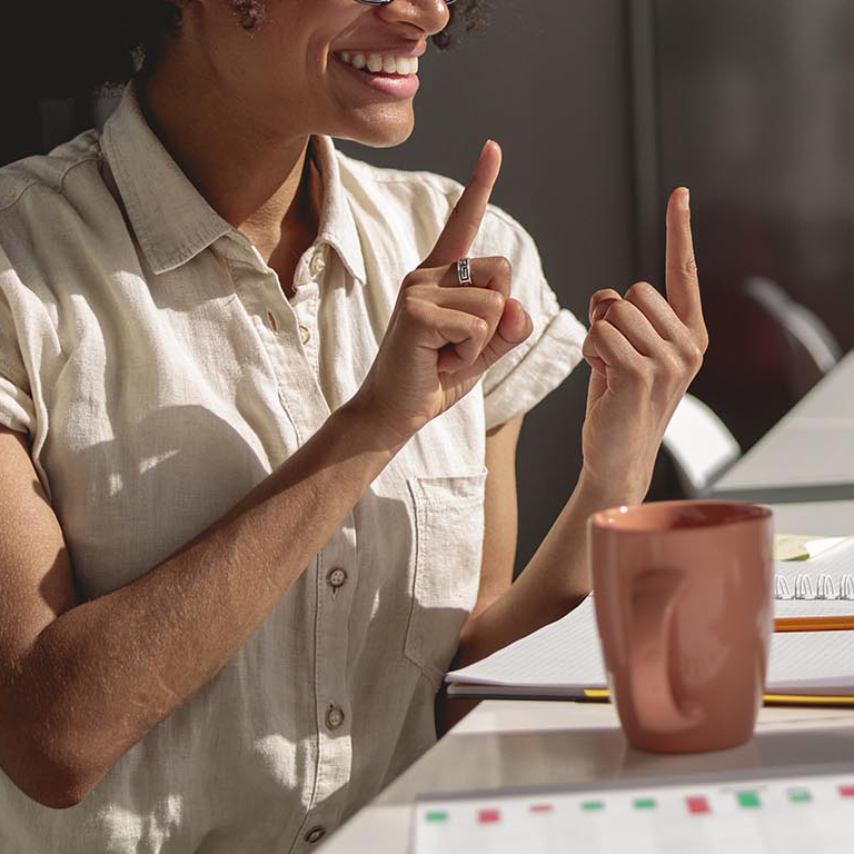 A female employee is at a desk communicating with a colleague through a virtual meeting app and using sign language.