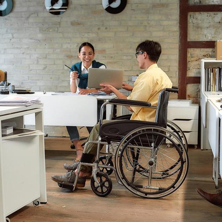 Office setting with disabled male employee in wheelchair presenting to female co-worker.