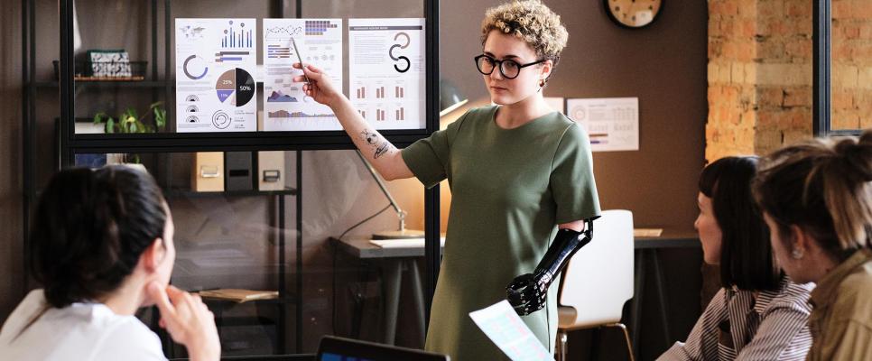 Woman with a prosthetic arm standing before seated group gestures at chart